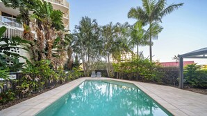 A rectangular swimming pool with a tiled border, surrounded by lush greenery and a building with balconies. - Coastal Rooftop Escape Mooloolaba - Walk to Beach (Mooloolaba Region)