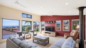 Ein modernes Wohnzimmer mit einer großen Couch, einem Couchtisch und einem Fernseher. Große Fenster bieten einen Blick ins Freie.