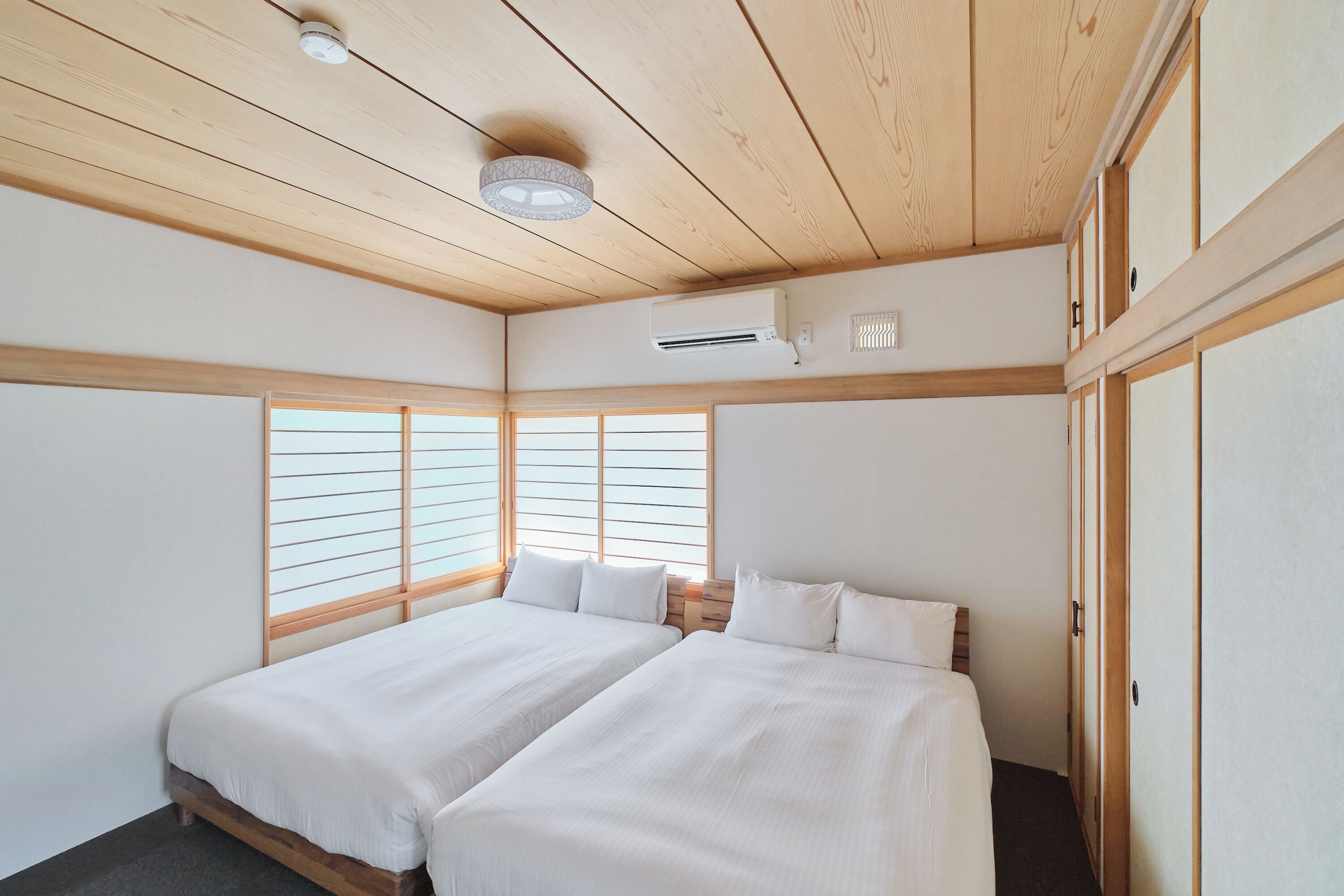 A room with two beds, wooden ceiling beams, an air conditioning unit, and large windows with horizontal blinds.