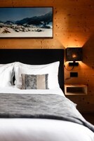 Cama com roupa de cama branca e cinza, cabeceira de madeira e mesa de cabeceira fixada na parede com uma luminária.