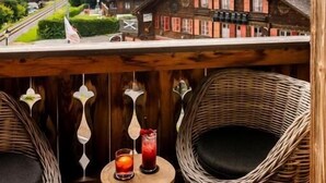 A wooden balcony with wicker chairs and a table, overlooking a village and mountains.