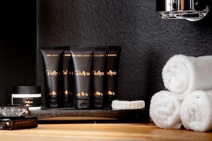 A set of black toiletry bottles with gold lettering, a small jar of cream, and rolled white towels on a wooden surface.