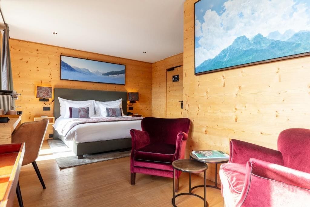 Une pièce aux murs lambrissés, avec un lit, un fauteuil rouge, une petite table et un tableau représentant des montagnes.