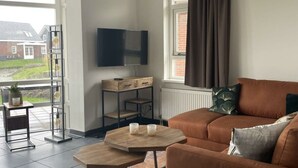 Un salon avec un canapé marron, un téléviseur fixé au mur, une fenêtre avec des rideaux et une plante posée sur une table d’appoint.