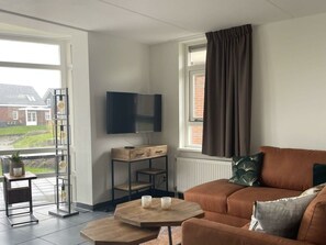 Ein Wohnzimmer mit einem braunen Sofa, einem an der Wand befestigten Fernseher, einem Fenster mit Vorhängen und einer Pflanze auf einem Beistelltisch.