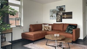 Un salon moderne avec un canapé d’angle marron, une table basse ronde et une grande fenêtre donnant sur un immeuble en briques.