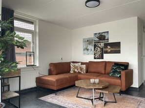Ein modernes Wohnzimmer mit einem braunen Ecksofa, einem runden Couchtisch und einem großen Fenster mit Blick auf ein Ziegelgebäude.