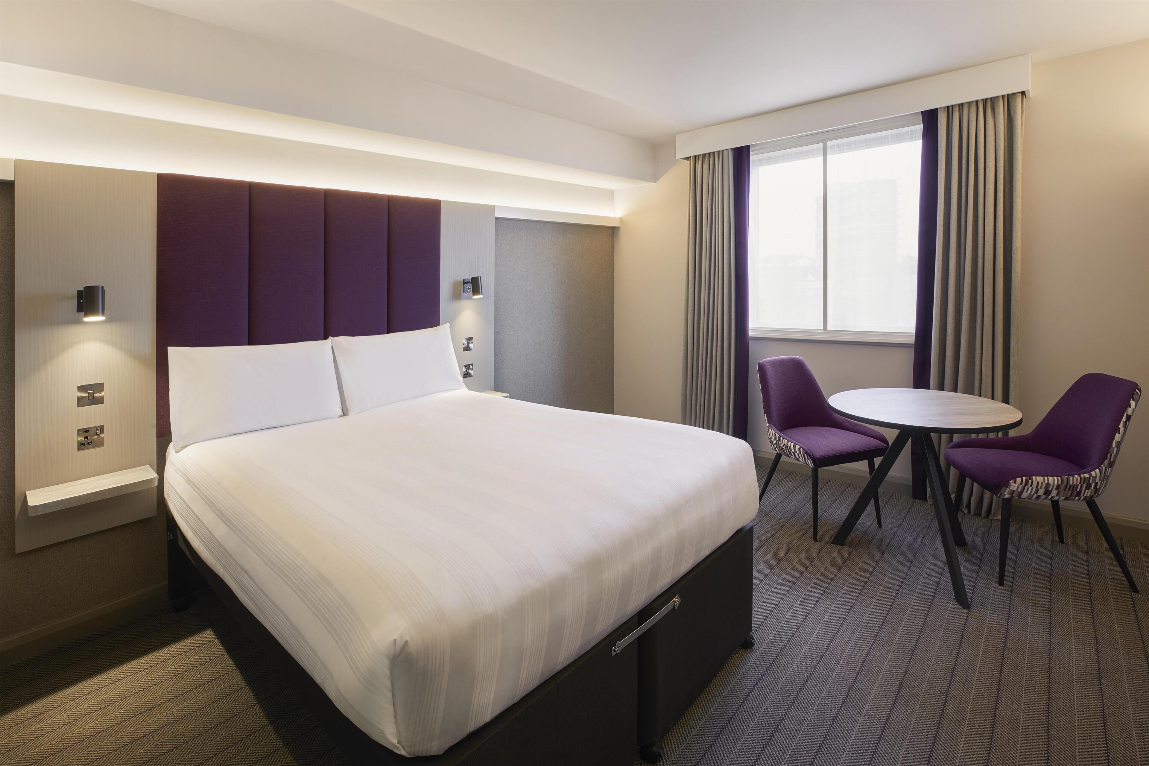 A hotel room with a bed, bedside tables, a desk, two chairs, and a window with curtains.