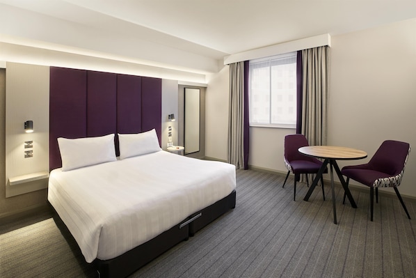 Una habitación de hotel con una cama grande, una mesa redonda, dos sillas, un espejo y una ventana con cortinas.
