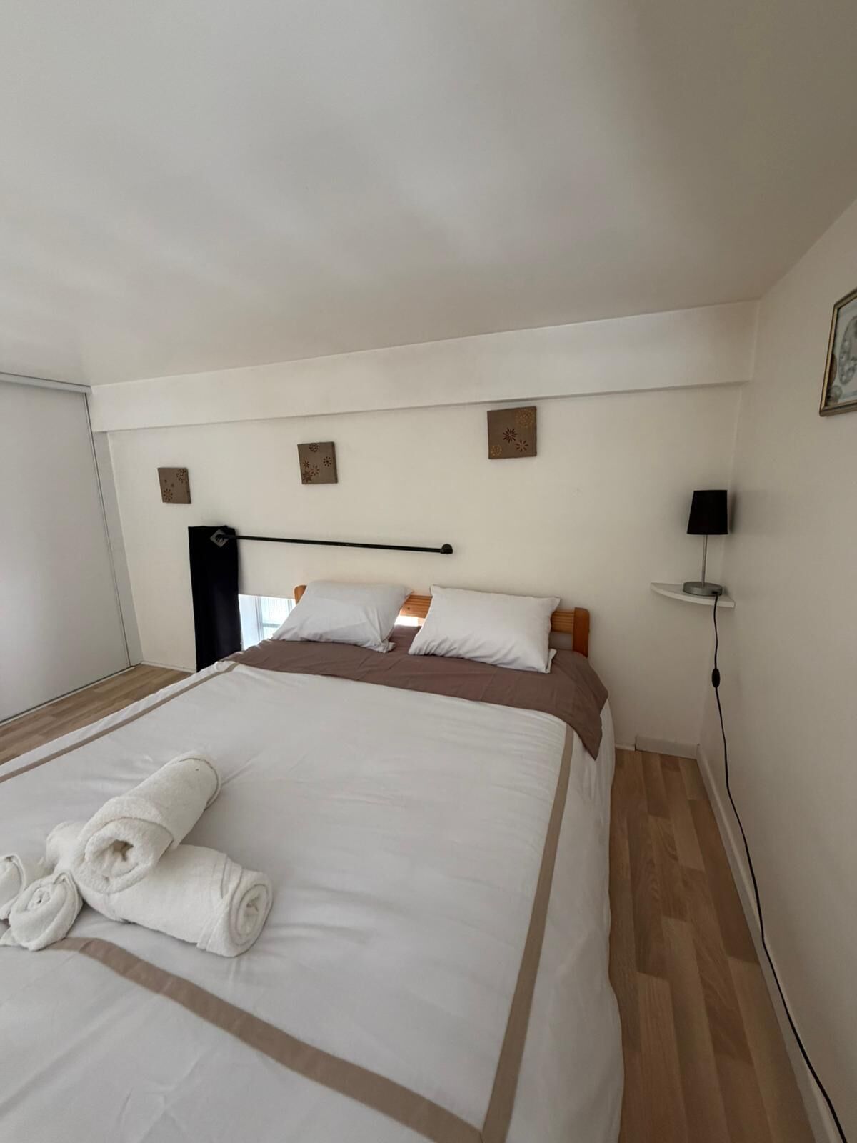 A neatly made bed with rolled towels, a black headboard, a wall-mounted lamp, and a small shelf.