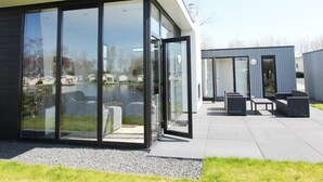 Un bâtiment moderne doté de grandes fenêtres en verre, d’une terrasse en béton et d’un espace extérieur aménagé pour s’asseoir.