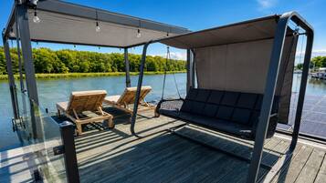 A covered outdoor seating area with two wooden lounge chairs and a swing seat, overlooking a calm body of water and greenery.