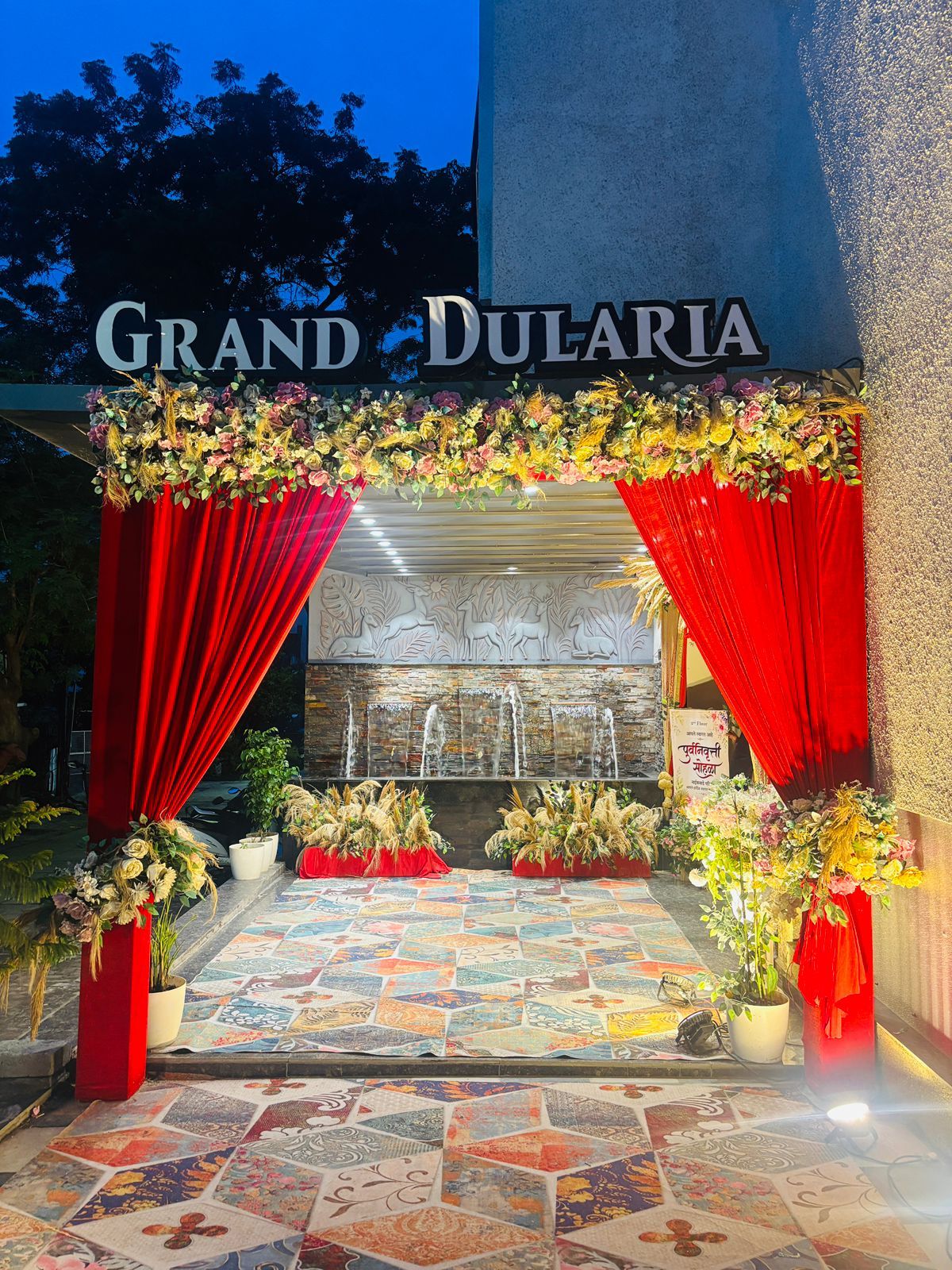 Una entrada con una cortina roja, decoraciones florales y un letrero que dice 'GRAND DULARIA'.