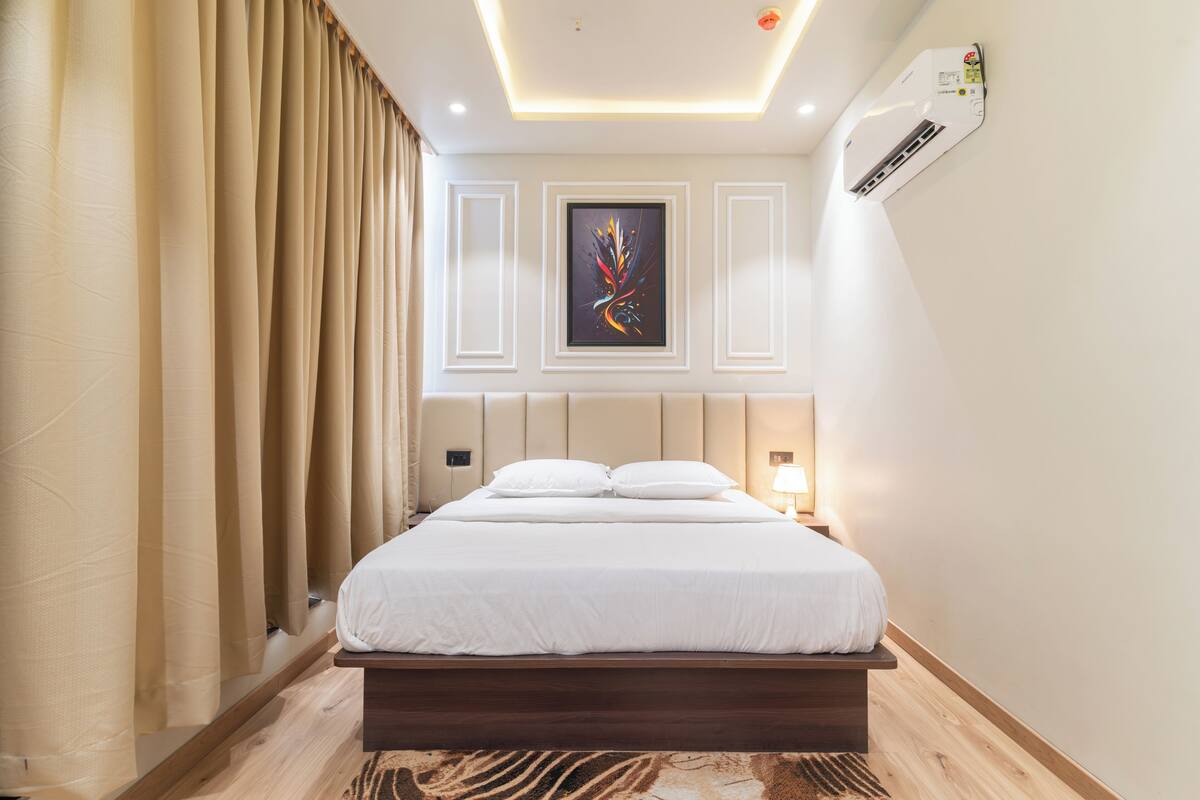 A hotel room with a bed, curtains, a wall-mounted air conditioner, and a framed artwork.