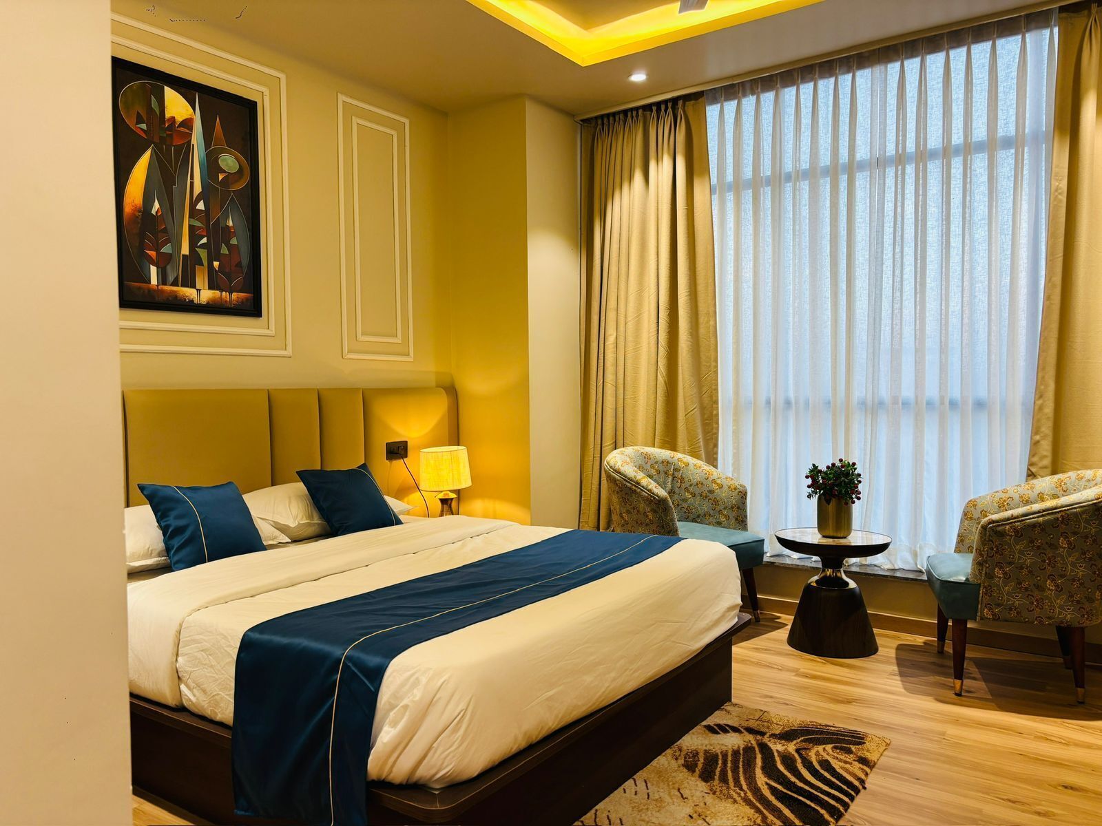 Una habitación de hotel con una cama grande, un escritorio con lámpara, dos sillas, una ventana con cortinas y un cuadro en la pared.