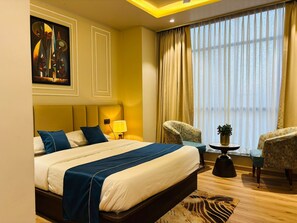 A hotel room with a large bed, a desk with a lamp, two chairs, a window with curtains, and a painting on the wall. - Grand Dularia (Nagpur)