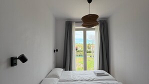 Ein Schlafzimmer mit Bett, Nachttisch, Pendelleuchte und Blick durch ein mit Vorhängen drapiertes Fenster.