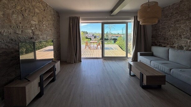 Ein modernes Wohnzimmer mit Steinwand, Flachbildfernseher, einer Couch, einem Couchtisch und Blick durch eine Schiebetür auf eine Terrassenanlage.