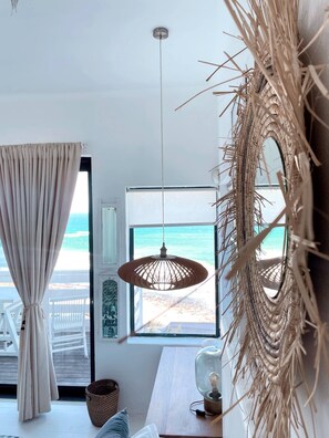 Een kamer met een groot raam met uitzicht op het strand, een geweven wanddecoratie, een hanglamp en een houten tafel.