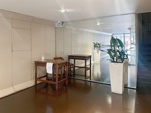 Een ruime kamer met een houten tafel en stoelen, een witte vaas met een plant, en een groot raam dat voor natuurlijk licht zorgt.