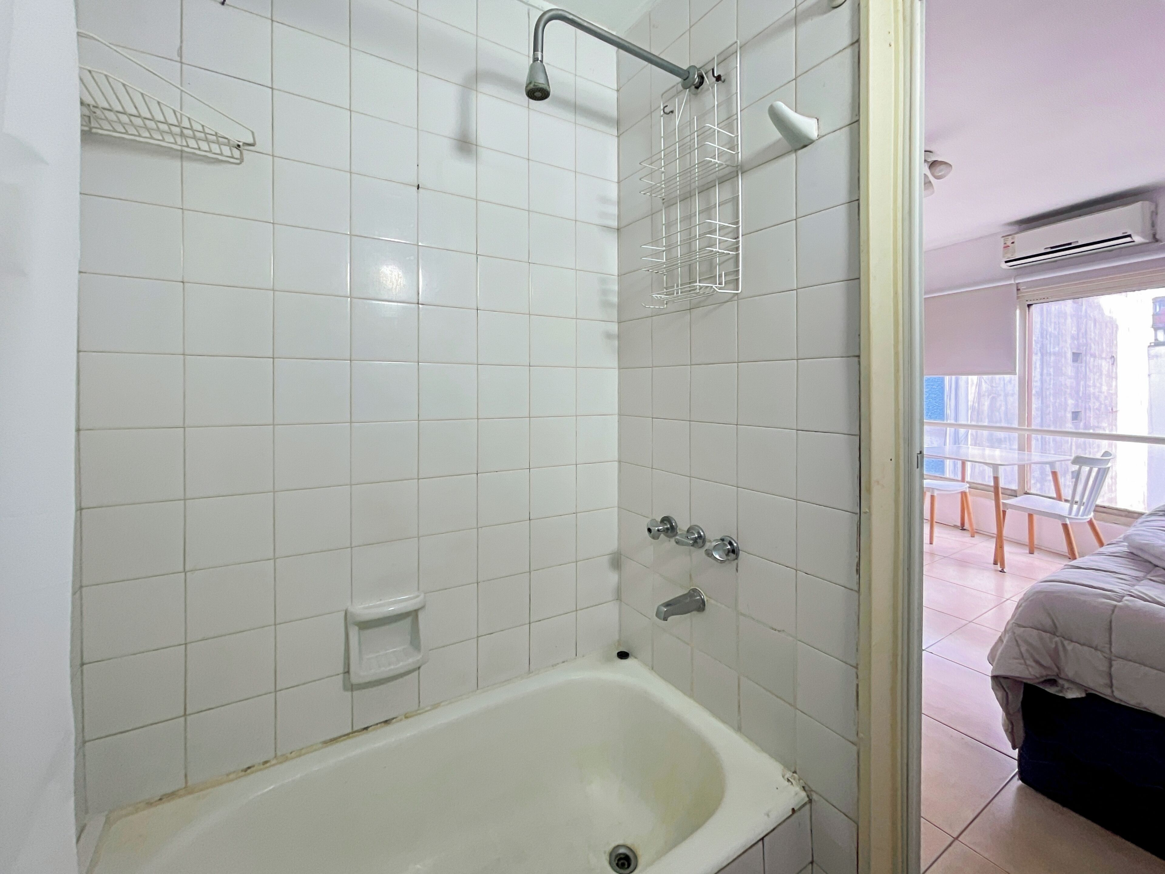 Ein Badezimmer mit weißen Fliesen, einer Badewanne, einer Dusche und einem Fenster mit Balkonblick.