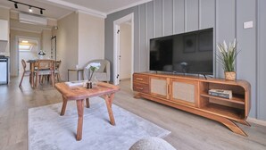 A living room with a wooden TV stand, a flat-screen TV, and a dining area with a wicker chair and table. - Refined 2BR with Terrace and Bosphorus View (Kadıköy)