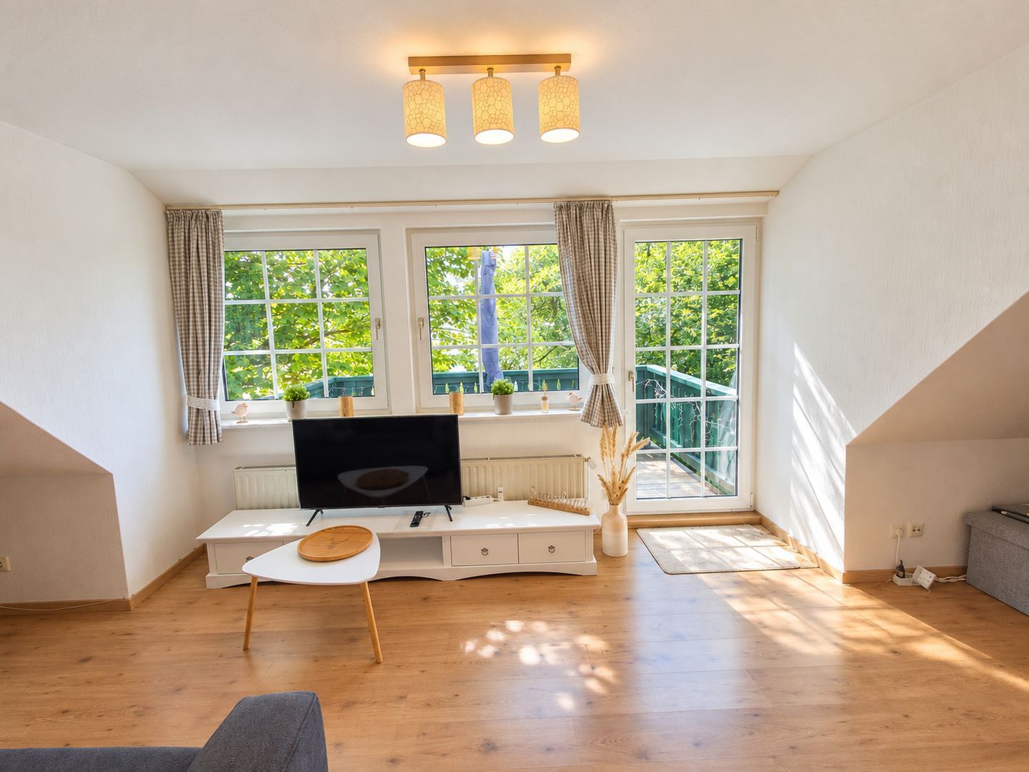 Een moderne woonkamer met een flatscreen televisie, een klein tafeltje en een bankje. Twee ramen met gordijnen, een trap en een uitzicht op groen.