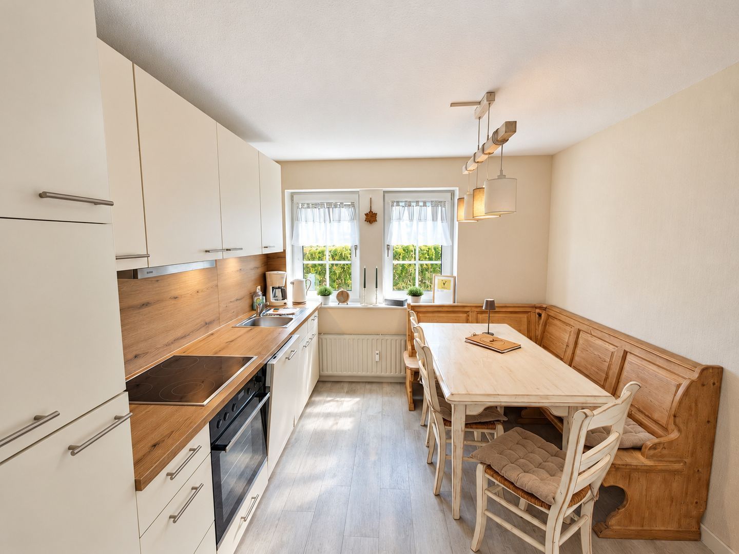 A modern kitchen with white cabinets, a wooden countertop, and a built-in oven. There is a window with curtains, a table with chairs, and a bench.