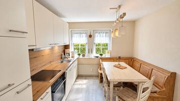 A modern kitchen with white cabinets, a wooden countertop, and a built-in oven. There is a window with curtains, a table with chairs, and a bench.
