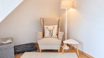 Una habitación de esquina con techo alto, un sillón beige con un cojín con motivos de ciervo, una mesita pequeña blanca, una lámpara de pie y un sofá gris, además de una alfombra blanca.