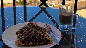 Een tafel met een bord wafel en een glas koffie, met uitzicht op een strand met parasols en palmbomen.