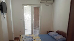 A bedroom with a bed, a desk, a chair, a window with blinds, an air conditioning unit, and a television on the wall. - Trokrevetni partman sa svojim kupatilom, terasom i parkingom u dvoristu kuce. (Budva)