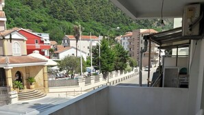 A balcony with a view of a street and buildings, a small table and chairs, and a railing. - Trokrevetni partman sa svojim kupatilom, terasom i parkingom u dvoristu kuce. (Budva)