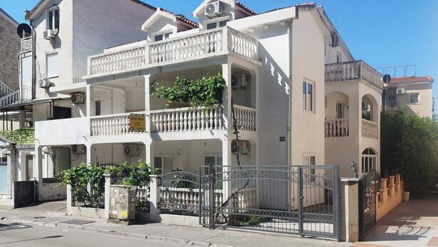 A multi-story building with balconies, a gated entrance, and a clear sky. - Trokrevetni partman sa svojim kupatilom, terasom i parkingom u dvoristu kuce. (Budva)