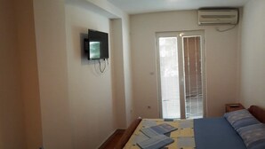 A bedroom with a bed, a TV mounted on the wall, a window with blinds, and an air conditioning unit. - Trokrevetni partman sa svojim kupatilom, terasom i parkingom u dvoristu kuce. (Budva)