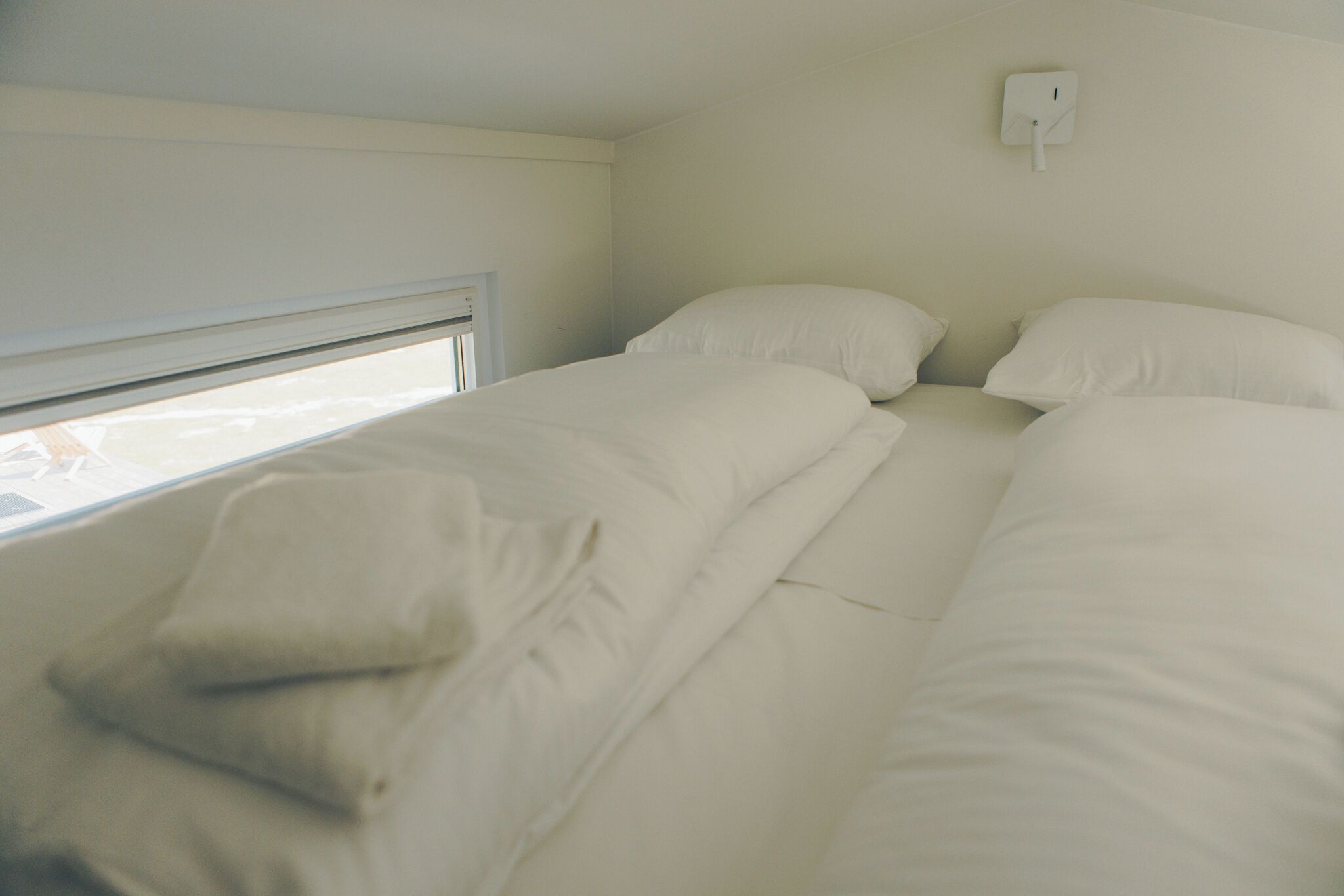 A single bed with white bedding in a room with a window and a wall-mounted light switch.