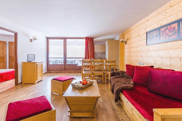 Een gezellige kamer met houten vloer, een rode bank, een houten tafel met fruit, een houten bankje en uitzicht op een balkon met een tafel en stoelen.