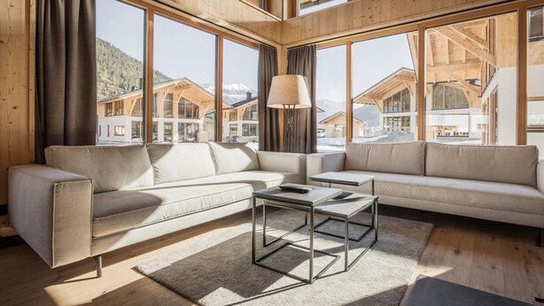 Un interior modern amb un sofà modular, una taula de centre i grans finestres que ofereixen vistes a cases de muntanya.