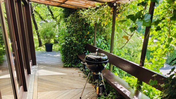 Una terraza de madera con una parrilla, rodeada de follaje y un banco de madera.