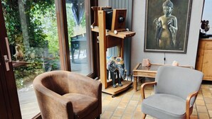 Una habitación con un sillón marrón, un sillón gris, una mesa de vidrio con objetos decorativos, un estante de madera con una cámara y una pintura de Buda.