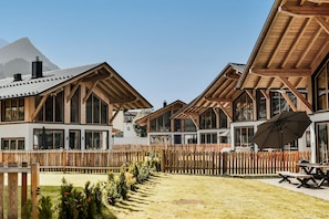 A row of wooden chalet-style buildings with large windows, a wooden fence, and a grassy area with outdoor seating.