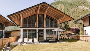 Een modern huis met een ruim terras, houten balken en een berg op de achtergrond.