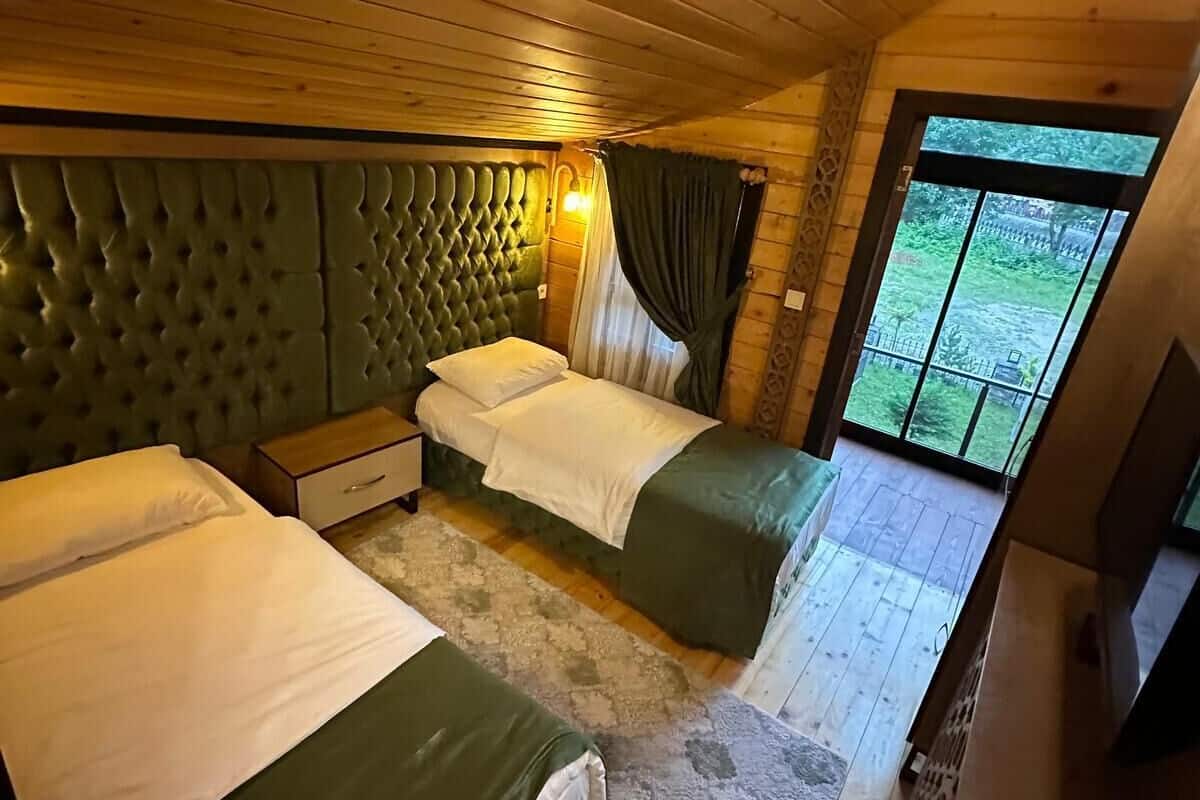 A cabin with two beds, a wooden ceiling, a window with curtains, and a view of greenery outside.