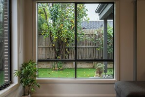 A room with a large window overlooking a backyard with a wooden fence, a tree, and a potted plant. - Seabreeze at Oakdean, Pet-Friendly (Ocean Grove)