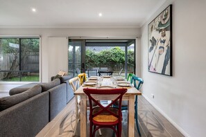 A dining area with a table set for four, a sofa, and a large window with a view of greenery. - Seabreeze at Oakdean, Pet-Friendly (Ocean Grove)