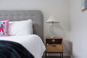 Une chambre à coucher avec une tête de lit rembourrée de couleur grise, un couvre-lit blanc, un coussin décoratif, une table de chevet avec une lampe et une petite table de nuit en bois.