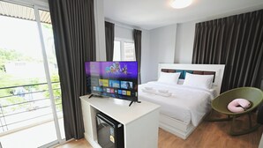 A hotel room with a bed, a TV on a stand, a small refrigerator, a chair, and a balcony with curtains.