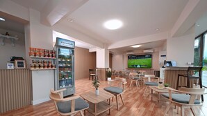 A modern hotel lobby with wooden flooring, a refrigerated beverage station, seating area, and a large TV screen displaying a sports game.