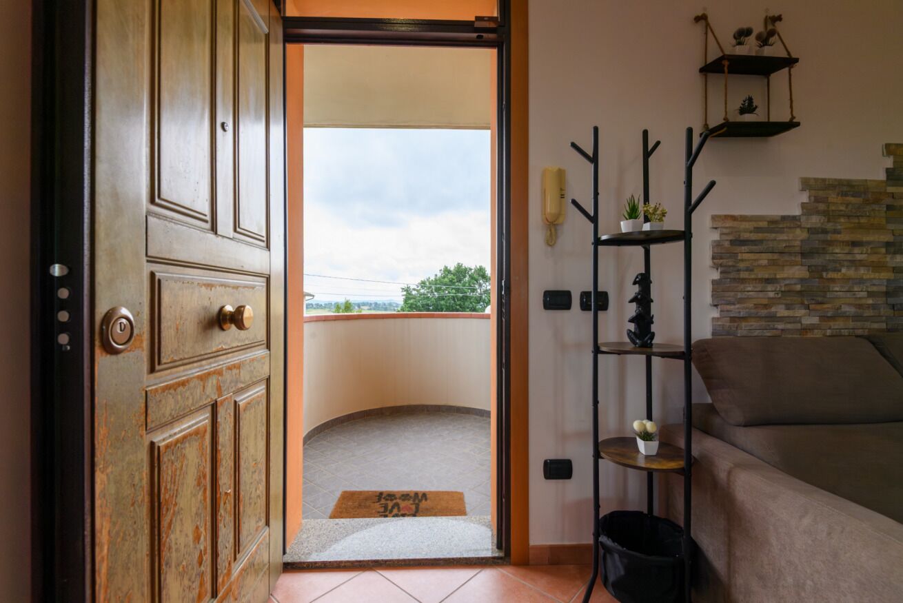 A room with a wooden door, a balcony with a doormat, a black metal shelf with plants, and a stone wall.