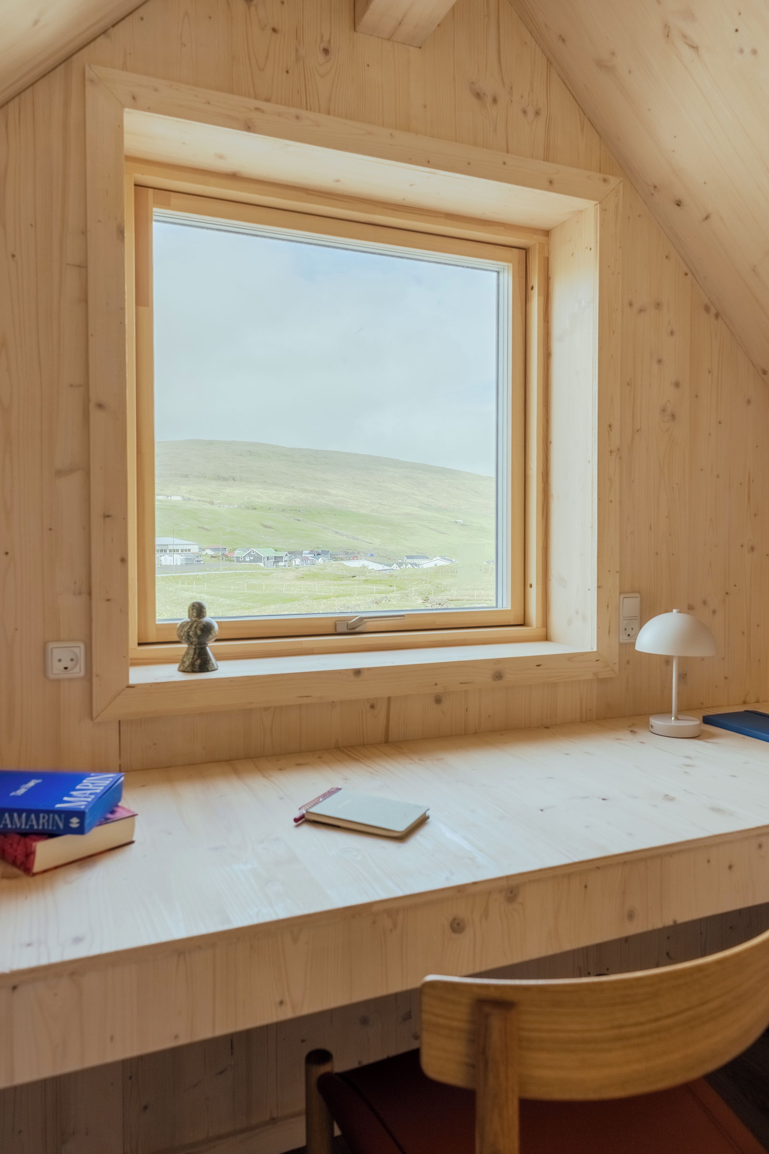 Una habitación con un escritorio de madera, una silla, un ventanal con vistas a un paisaje verde y una lámpara.